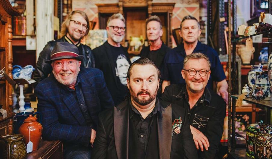 A group of seven men pose together in an antique store, surrounded by shelves filled with various vintage items like ceramics and teapots. The man in