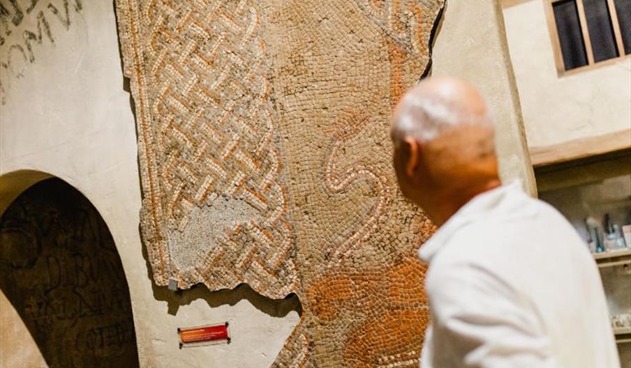 A man with short gray hair examines an ancient Roman mosaic with intricate geometric patterns displayed on a wall in the Hull & East Riding Museum of
