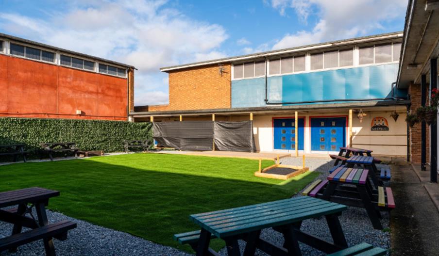 A courtyard with a neatly trimmed grassy area, surrounded by gravel paths and several colorful wooden picnic tables. The buildings around the courtyar