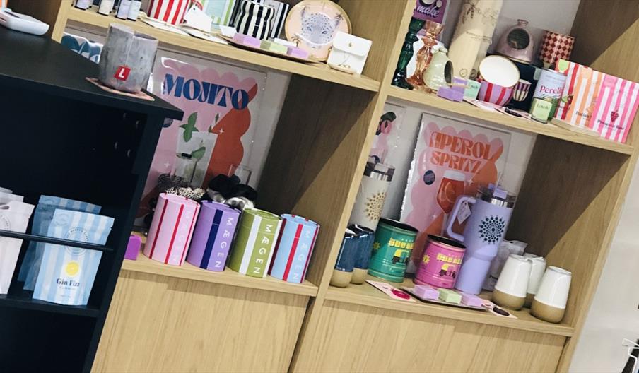 Colorful shop display with wooden shelves filled with gift items. The shelves hold striped bowls, small candle holders, mugs, tins in pastel colors la