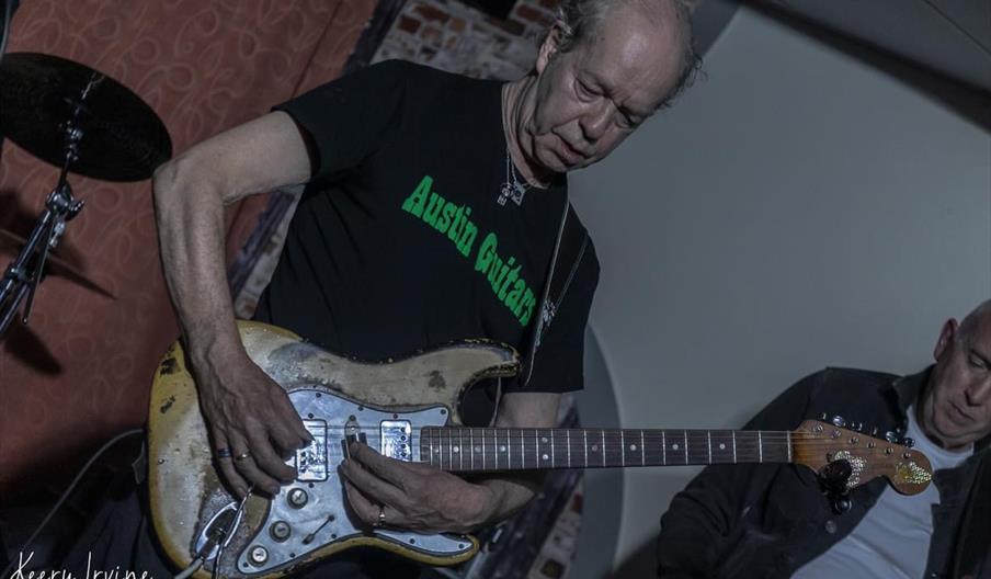 An older man wearing a black "Austin Guitars" t-shirt plays an aged electric guitar on stage, with another musician partially visible beside him and a