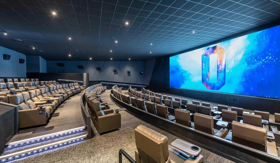 The inside of a modern cinema, with a large colourful screen and rows of beige recliner chairs facing it.
