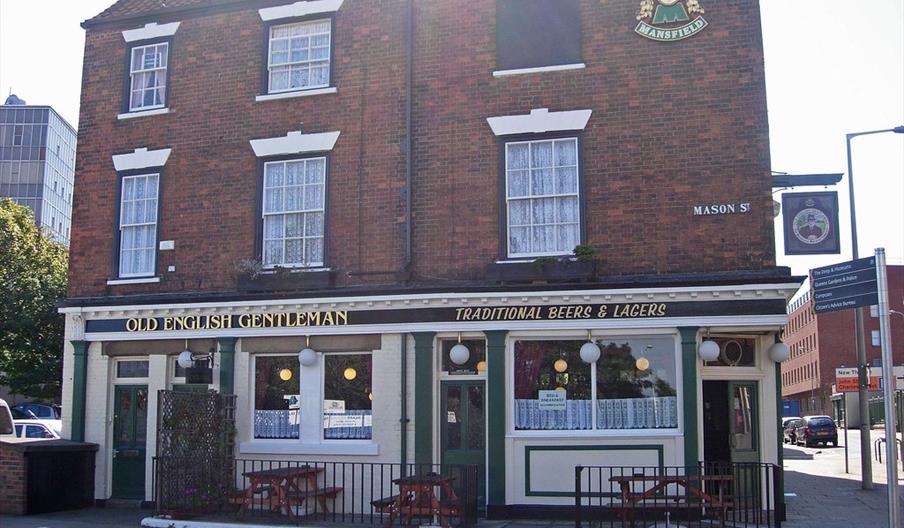 A traditional English pub called "Old English Gentleman" with a brick exterior, white-trimmed windows, and lace curtains. Outdoor seating with wooden