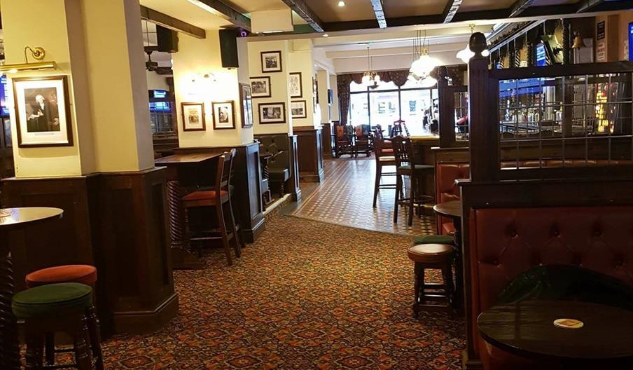 Inside of pub with wood and glass partition, dark wood stools, chairs and tables with red and green floral carpet.