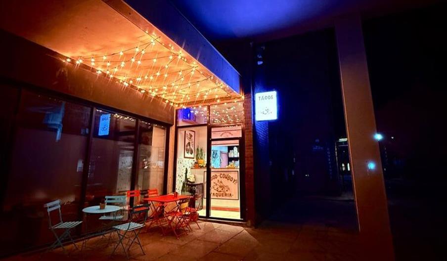 Exterior of Food For Cowboys at night with  string lights hanging above its entrance, outdoor tables and chairs arranged on the sidewalk, and a glowin