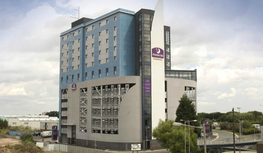 A modern multi-story Premier Inn hotel building with distinctive blue and gray exterior panels, prominent purple signage, and adjacent roadways, set a