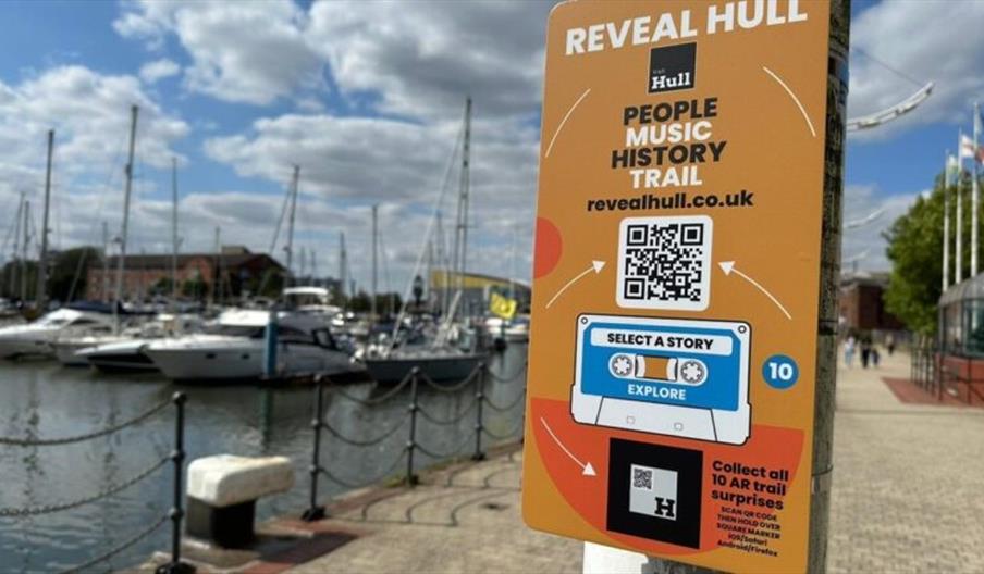 A sign reading 'Reveal Hull' along with a QR code is attached to a lamppost, with a marina filled with boats in the background.