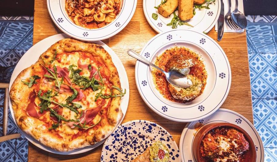 A wooden table set with various Italian dishes, including a pizza topped with arugula and prosciutto, a pasta dish, breaded fried sticks with greens,