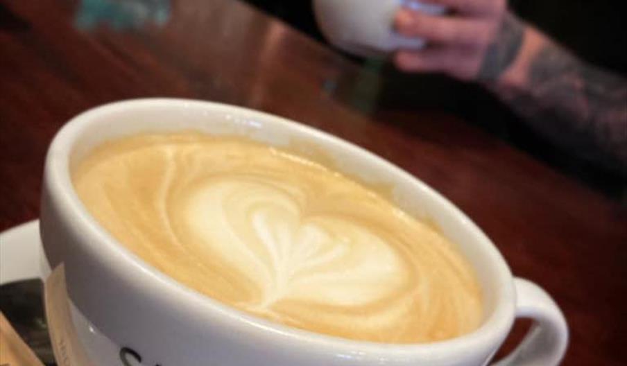 A close-up of a cup of latte with heart-shaped latte art on top, served in a white cup labeled "SLEEPERS CAFE BAR" on a saucer with a brown sugar pack
