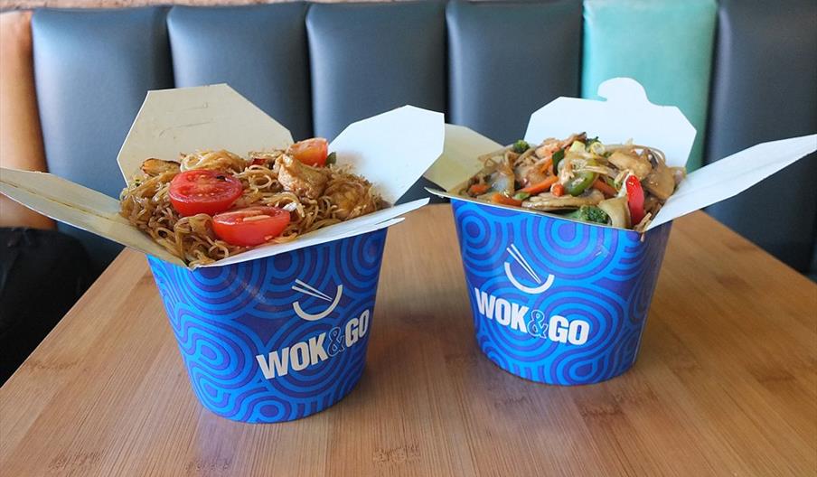 Two blue Wok and Go branded takeout boxes filled with stir fry and noodles on a wooden table.
