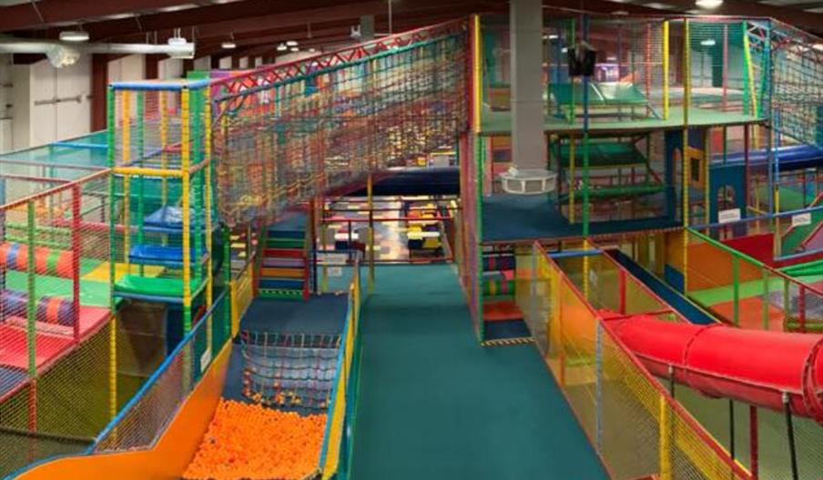 A soft play area for children with multicoloured netting, an enclosed slide, an open slide into a ballpool below and stairs leading upwards.