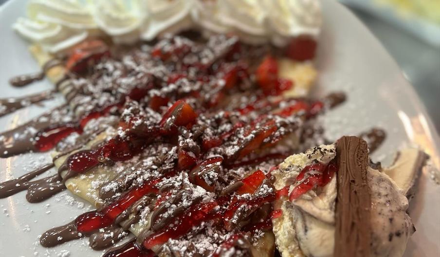 A plated dessert featuring a folded crepe topped with chocolate and strawberry sauce, powdered sugar, and sliced strawberries, served with a scoop of