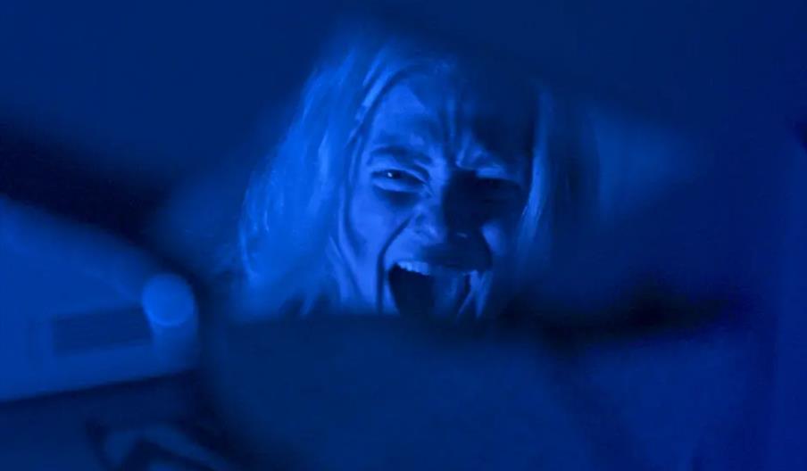 A person with long hair is shown screaming or yelling, their face illuminated by blue light and reflected in a fragmented mirror. The image has a dark