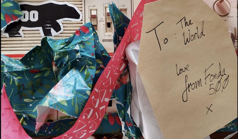 A partially unwrapped gift with blue and green festive wrapping paper, a pink ribbon, and a large beige gift tag. The tag reads "To: The World, love f