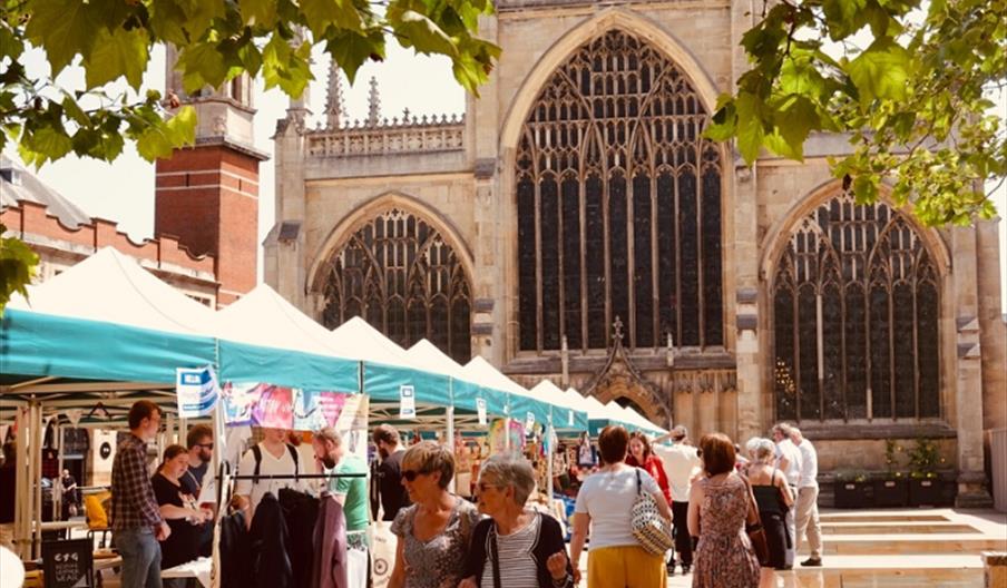 An outdoor market in Trinity Square with green and white tents is set up in front of a large, ornate gothic-style church, with people browsing stalls