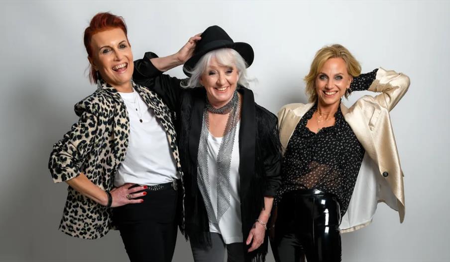 Three women stand together in stylish outfits, smiling and posing confidently against a plain white background. The woman on the left wears a leopard
