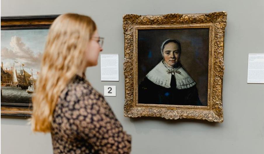 A woman with long blonde hair stands closely observing a framed portrait of a woman in historical dress at the Ferens Art Gallery. The portrait is hun