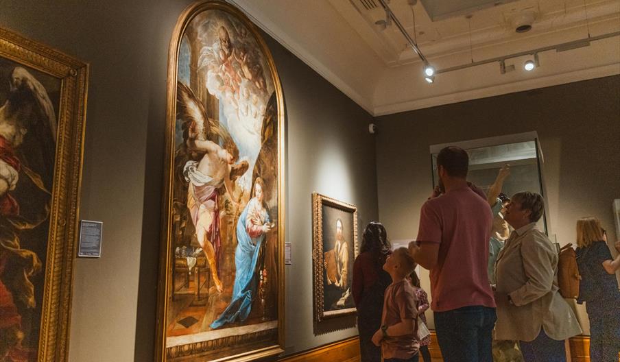 A group of people stand in a museum gallery looking at large, framed classical paintings on the walls. The room is softly lit and some visitors are ob
