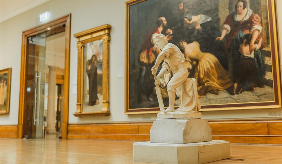 A marble sculpture of a young boy holding a cricket bat is displayed on a pedestal in the Ferens Art Gallery, with large framed paintings featuring fi