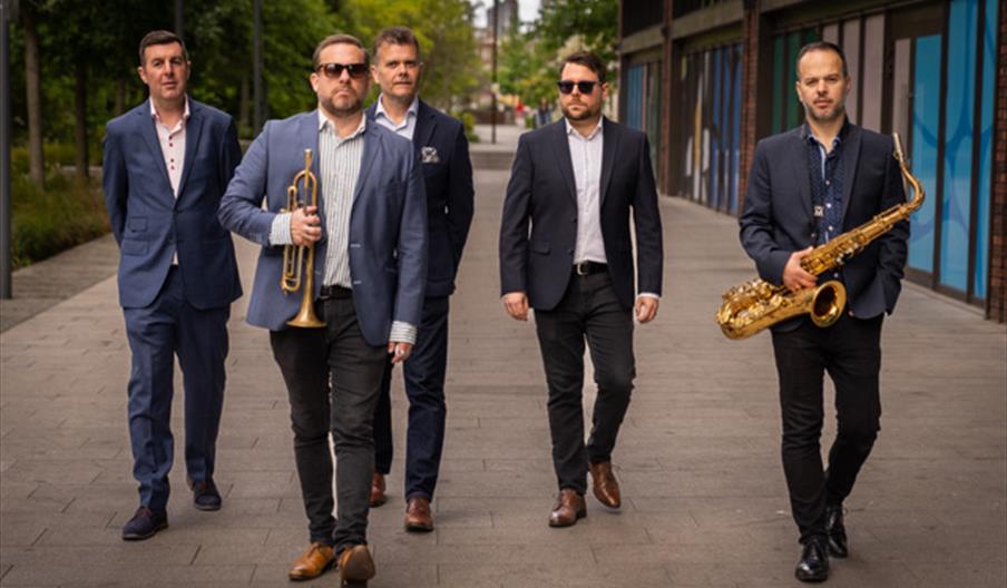Five men in suits walk down a city sidewalk, with one holding a trumpet and another holding a saxophone, suggesting they are musicians. Trees and mode
