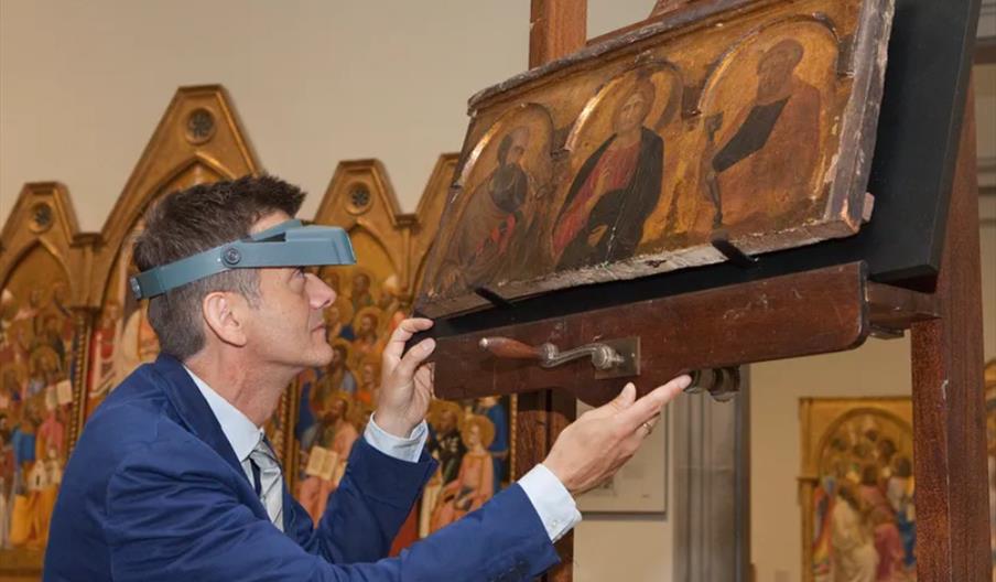 A conservator wearing magnifying headgear examines a medieval religious painting on an easel at the Ferens Art Gallery, with other religious artworks