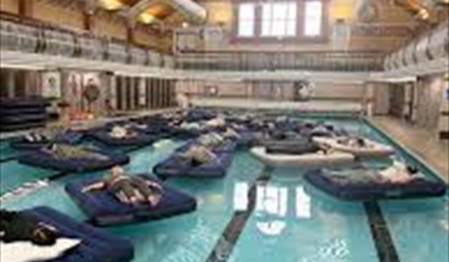 People are lying on air mattresses floating in an indoor swimming pool under a large glass ceiling in a spacious, well-lit building.
