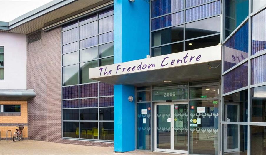 Exterior view of The Freedom Centre, a modern building with large glass windows and a blue pillar at the entrance; a sign above the door displays the