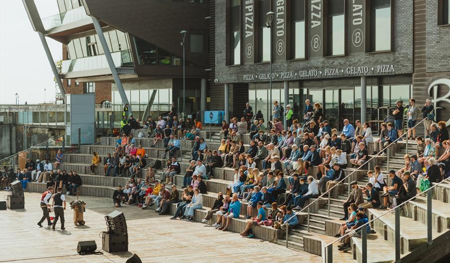 A large audience sits on tiered wooden steps outdoors, attentively watching two performers on a stage area at Stage@thedock in front of a modern build