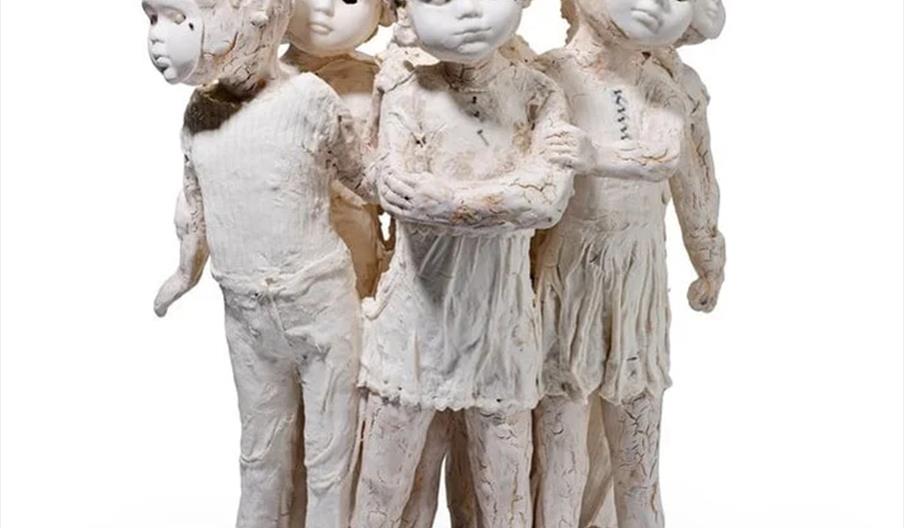 A ceramic sculpture depicting six children standing closely together in a cluster, all with white, textured surfaces and somber facial expressions. Th