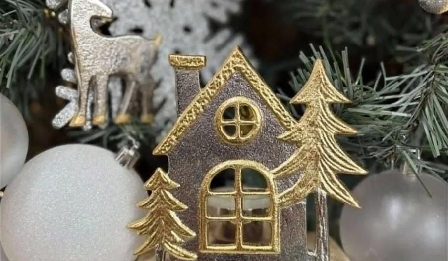 A metallic ornament shaped like a house with golden trees is displayed among pine branches, white baubles, and a silver reindeer decoration, creating