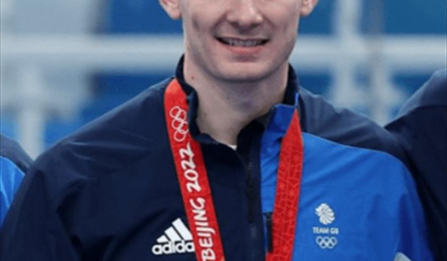 Grant Hardie wearing a blue Team GB jacket smiles while holding a Beijing 2022 Olympic medal around his neck and a small bouquet of flowers.
