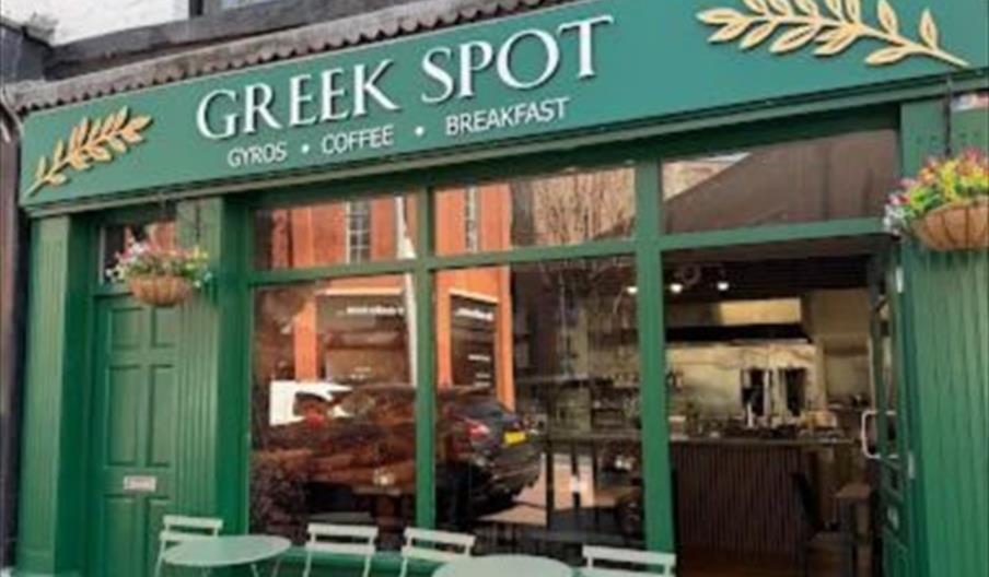 A small restaurant called "Greek Spot" with a green exterior, large windows, and a sign advertising gyros, coffee, and breakfast. There are green tabl