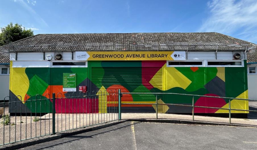 The front of Greenwood Avenue Library, a single-story building with a mural of bold, colorful geometric shapes covering its metal shutters and walls,