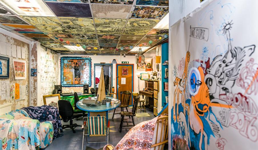 A colourful, eclectic room at Ground Galley filled with mismatched chairs, couches draped with patterned blankets, and a round table at the center. Th