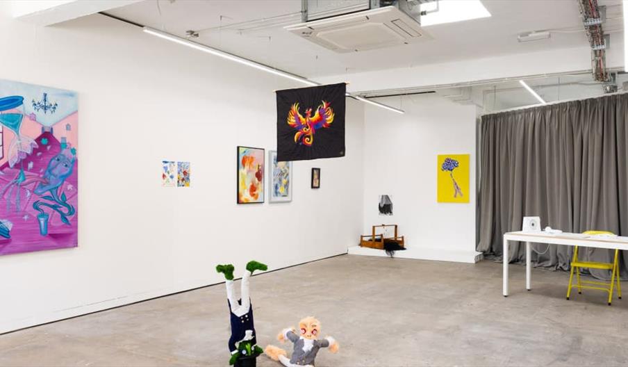 Contemporary art gallery interior with abstract paintings on white walls, a hanging black textile art piece with a colorful phoenix, two plush sculptu