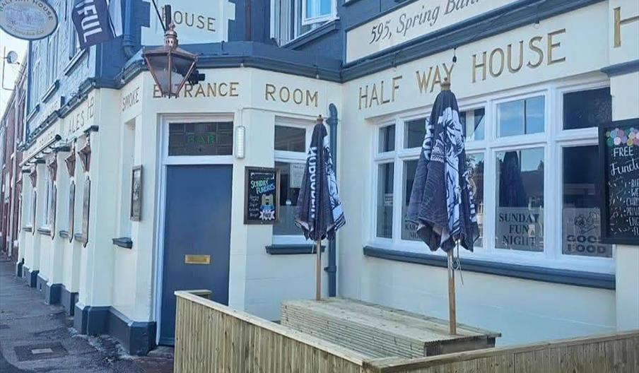 Corner view of the Half Way House pub with cream and blue exterior, signage displaying the address "595, Spring Bank West," and a small wooden outdoor
