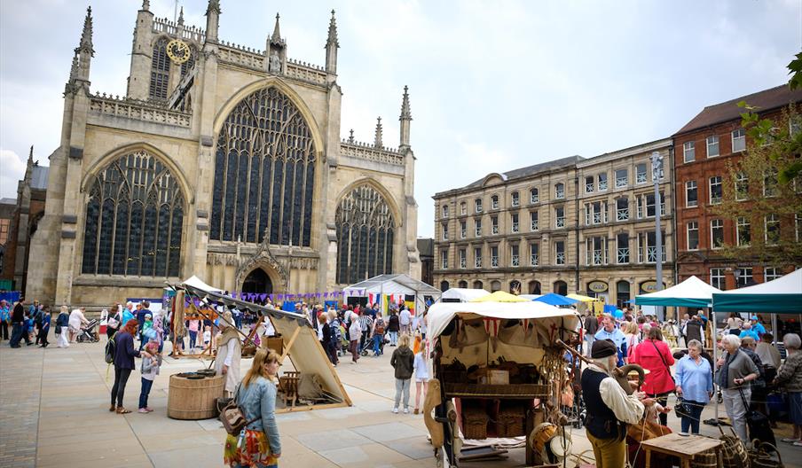 A bustling outdoor market with people mingling among stalls and tents, set in front of a large Gothic-style church with tall arched windows and decora