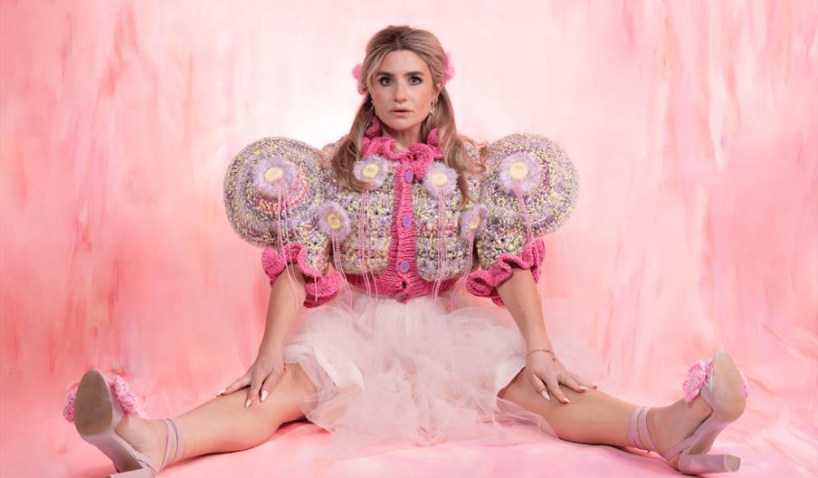 A woman sits on the floor with her legs stretched out and arms resting on her knees, wearing a whimsical pink and purple knitted top with exaggerated