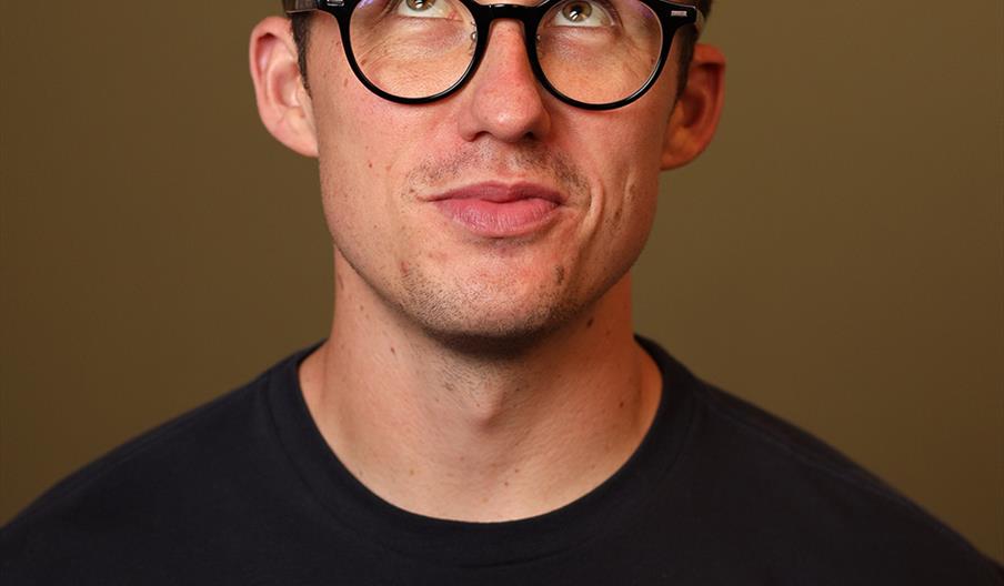 A man wearing black glasses and a dark t-shirt looks upward with a thoughtful expression against a plain background.