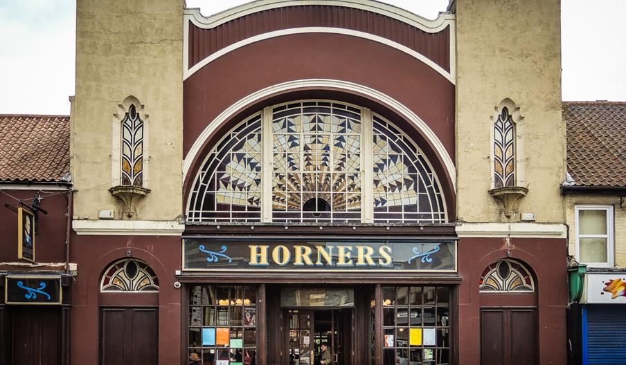 Front view of an old-fashioned pub called "Horners" with a large arched stained glass window above the entrance, flanked by beige towers and decorativ