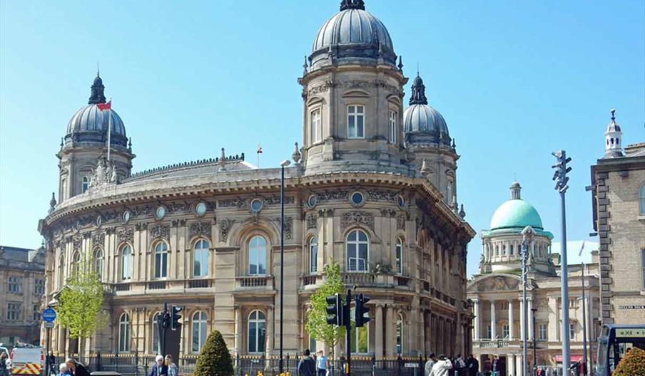 An external shot of the Hull Maritime museum, a building with multiple towers and ornate architecture. Below are people wandering across a grassy verg