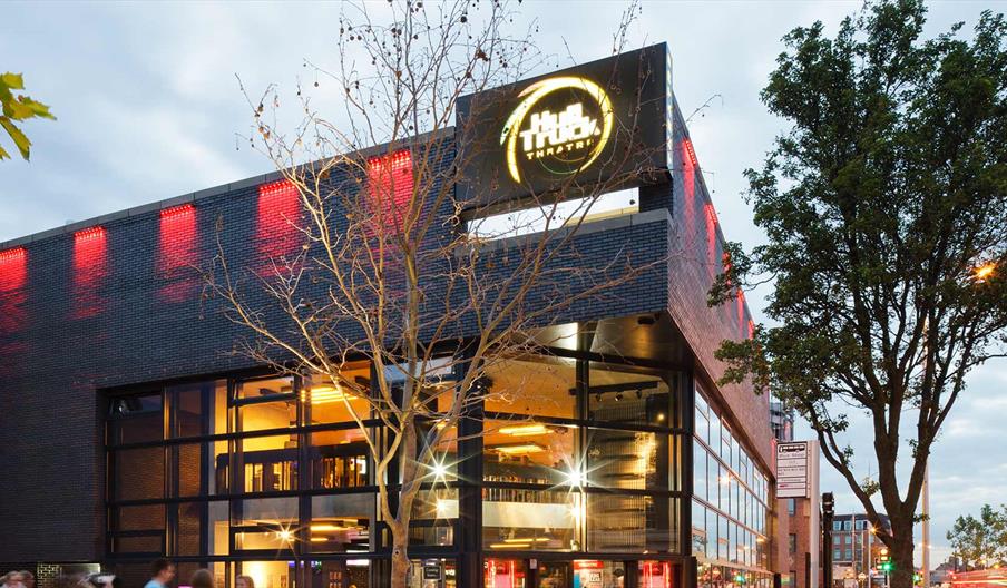 A modern theater building with large glass windows, illuminated by warm interior lights and red exterior lights, with a prominent sign reading "Hull T