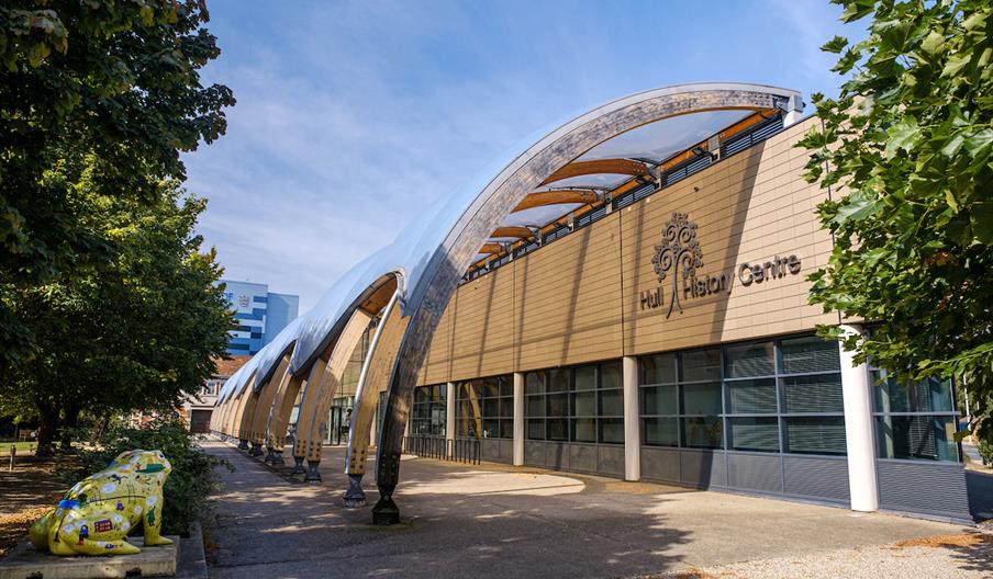 Modern building with large, curved arches and glass windows, labeled "Hull History Centre," surrounded by trees and featuring a brightly painted frog