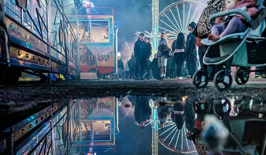 A night-time carnival scene shows crowds of people, colourful amusement rides, and bright lights, all reflected clearly in a large puddle on the groun