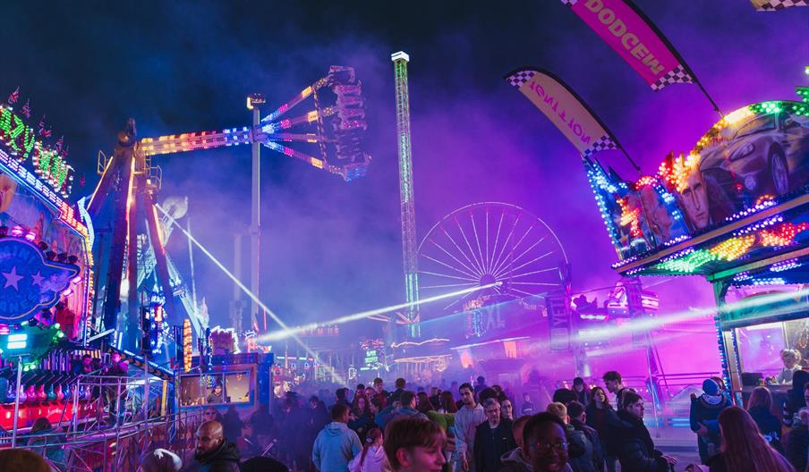 A lively nighttime carnival scene with a large crowd of people walking among bright, colorful amusement rides and attractions, including a Ferris whee
