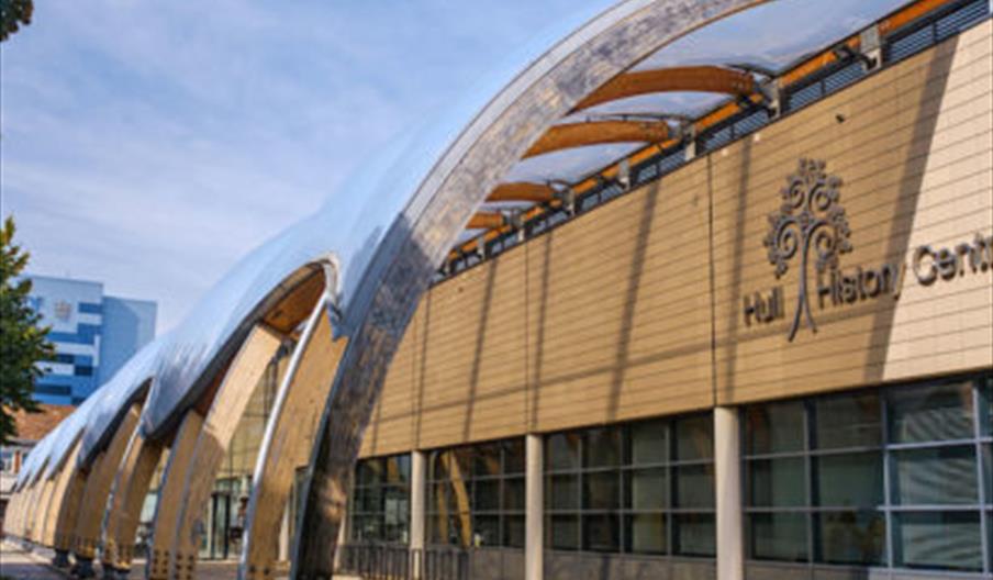 The image shows the exterior of the Hull History Centre, featuring a modern building with large windows and distinctive curved metal arches along the