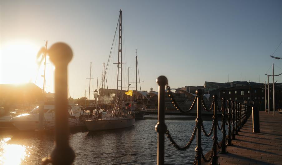 Hull marina at sunset with several docked sailboats, water reflecting the sunlight, and a walkway lined with chain-linked posts on the right side of t
