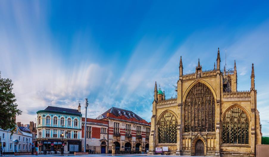 Hull Minster, a large Gothic-style church with tall spires and arched windows stands beside older buildings, all reflected in a shallow pool of water