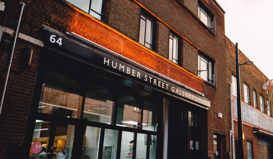 The image shows the exterior of the Humber Street Gallery, a brick building with large windows and a glass entrance door. The gallery's name and stree