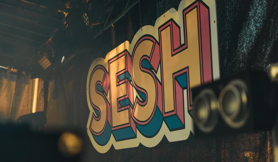 A large, colorful sign with the word "SESH" in bold, yellow letters outlined in pink and blue, hangs on a dark stage background with spotlights above.